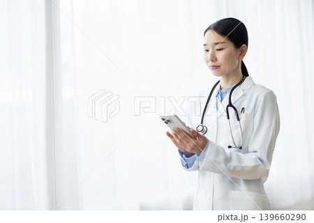 明るい窓辺でスマートフォンを操作する白衣を着た日本人女性医師 明るい窓辺でスマートフォンを操作する白衣を着た日本人女性医師 139660290