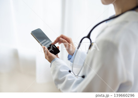 明るい窓辺でスマートフォンを操作する白衣を着た日本人女性医師の手元のクローズアップ 139660296