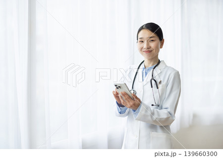 明るい窓辺でスマートフォンを操作する白衣を着た日本人女性医師 明るい窓辺でスマートフォンを操作する白衣を着た日本人女性医師 139660300