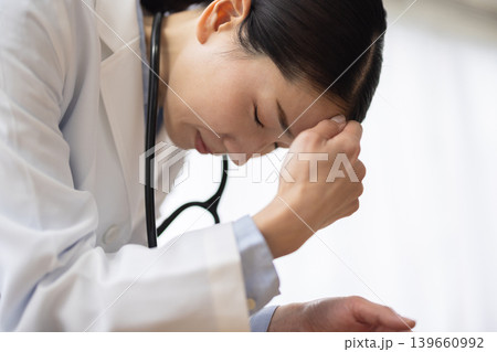 こめかみに触れて痛そうな表情をする白衣を着た日本人女性医師 こめかみに触れて痛そうな表情をする白衣を着た日本人女性医師 139660992