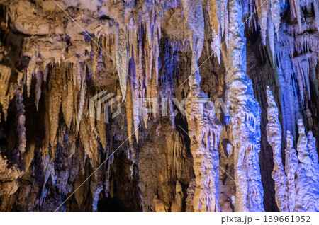 Dense sharp rock stalactite formations hang down from ancient underground limestone cave ceiling beautifully illuminated by contrasting warm and vibrant blue artificial lighting 139661052
