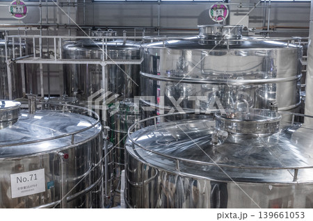 Close detailed view showing heavy reflective industrial stainless steel fermentation storage tanks standing inside spacious modern manufacturing facility processing liquid commercial beverage products Close detailed view showing heavy reflective industrial stainless steel fermentation storage tanks standing inside spacious modern manufacturing facility processing liquid commercial beverage products 139661053
