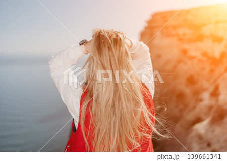 Woman sunset cliff standing on the edge overlooking sea holding long blonde hair expressing freedom during golden hour Woman sunset cliff standing on the edge overlooking sea holding long blonde hair expressing freedom during golden hour 139661341