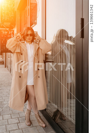 Fashion model street. Stylish woman in beige trench coat and sunglasses posing by glass building with warm sunlight Fashion model street. Stylish woman in beige trench coat and sunglasses posing by glass building with warm sunlight 139661342
