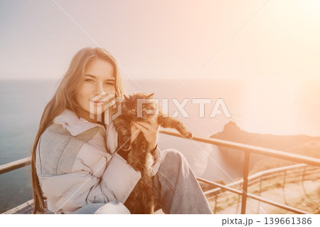 Woman cat sunset sitting at a scenic coastal viewpoint while holding her pet during a beautiful warm golden hour Woman cat sunset sitting at a scenic coastal viewpoint while holding her pet during a beautiful warm golden hour 139661386