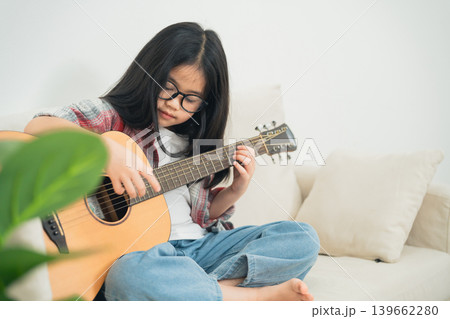 Young girl with glasses strumming an acoustic guitar while sitting on a couch in a cozy living room, showcasing her musical talent and creativity. Young girl with glasses strumming an acoustic guitar while sitting on a couch in a cozy living room, showcasing her musical talent and creativity. 139662280