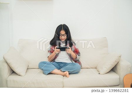 Young girl with glasses sitting on a couch playing a mobile game, wearing casual clothes, enjoying leisure time indoors with a relaxed atmosphere 139662281