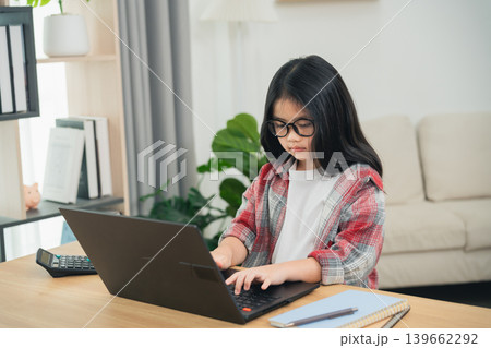 Young girl with glasses typing on laptop at home, focused on screen, studying or doing homework in cozy living room with plants and natural light Young girl with glasses typing on laptop at home, focused on screen, studying or doing homework in cozy living room with plants and natural light 139662292