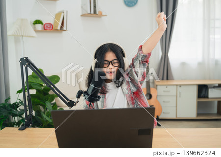 Young girl with glasses celebrating success while using a laptop in a cozy home office setting with a microphone and plants around her 139662324