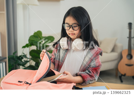Young girl with glasses and headphones preparing for school, looking inside pink backpack on desk in cozy room with guitar Young girl with glasses and headphones preparing for school, looking inside pink backpack on desk in cozy room with guitar 139662325