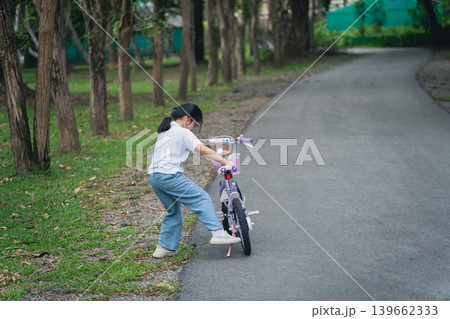 Young girl learning to ride a bicycle in a serene park environment, surrounded by trees and greenery, showcasing childhood innocence and adventure Young girl learning to ride a bicycle in a serene park environment, surrounded by trees and greenery, showcasing childhood innocence and adventure 139662333
