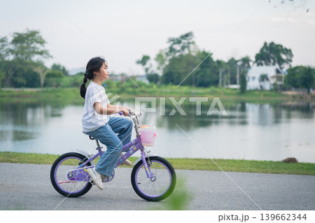 Happy girl riding a purple bicycle near a serene lake with lush greenery, enjoying a warm sunny day and embracing the joy of childhood and adventure 139662344