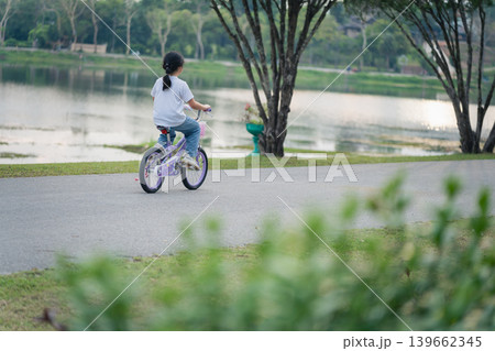 Child Riding Bicycle by Calm Lake in Park Surrounded by Lush Trees, Enjoying Outdoor Activity during Sunny Day with Clear Sky and Peaceful Atmosphere 139662345