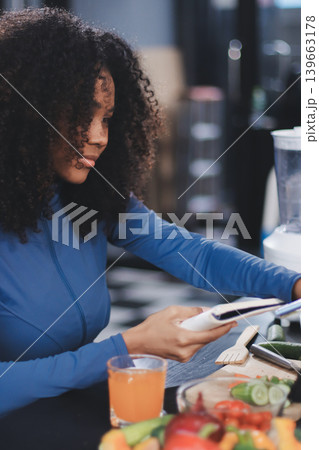Young nutritionist showing bell peppers during an online video call consulting about healthy eating, sitting at home at a table with vegetables, laptop and measuring tape 139663178