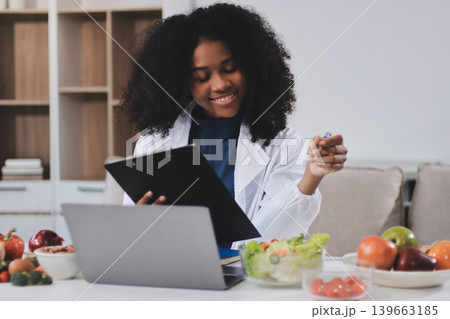 Young nutritionist showing bell peppers during an online video call consulting about healthy eating, sitting at home at a table with vegetables, laptop and measuring tape Young nutritionist showing bell peppers during an online video call consulting about healthy eating, sitting at home at a table with vegetables, laptop and measuring tape 139663185