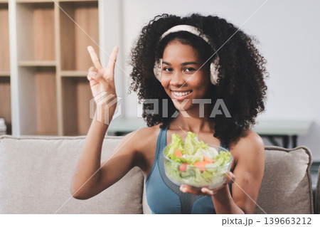 Young nutritionist showing bell peppers during an online video call consulting about healthy eating, sitting at home at a table with vegetables, laptop and measuring tape 139663212
