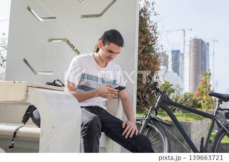 A young man is seated on a bench, looking at his phone with focus. A young man is seated on a bench, looking at his phone with focus. 139663721