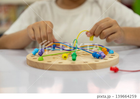 Close up of children's hands playing with a colorful wooden stringing toy, practicing fine motor skills and hand-eye coordination. Close up of children's hands playing with a colorful wooden stringing toy, practicing fine motor skills and hand-eye coordination. 139664259
