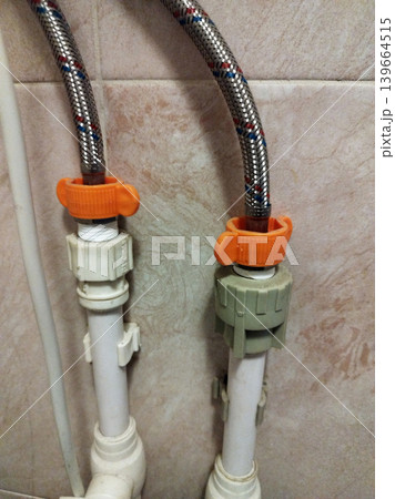 Flexible braided metal hoses are connected to white plastic pipes using orange butterfly-style wing nuts. This plumbing setup is mounted against a tiled wall, showing a typical home water installation Flexible braided metal hoses are connected to white plastic pipes using orange butterfly-style wing nuts. This plumbing setup is mounted against a tiled wall, showing a typical home water installation 139664515