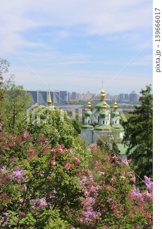 Green-domed church with golden spires is framed by blooming lilacs and a distant city skyline under a clear blue sky in Kyiv, showcasing springtime beauty Green-domed church with golden spires is framed by blooming lilacs and a distant city skyline under a clear blue sky in Kyiv, showcasing springtime beauty 139666017