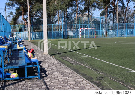 Soccer field with artificial turf featuring blue benches, colorful balls, and a goalpost surrounded by trees and a fence in a sports complex setting Soccer field with artificial turf featuring blue benches, colorful balls, and a goalpost surrounded by trees and a fence in a sports complex setting 139666022