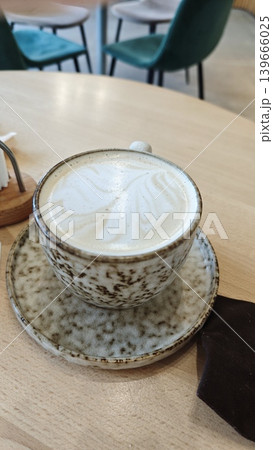 Latte art in a coffee cup on a saucer sits on a wooden table in a contemporary cafe, with blurred chairs and ambient lighting visible in the background Latte art in a coffee cup on a saucer sits on a wooden table in a contemporary cafe, with blurred chairs and ambient lighting visible in the background 139666025