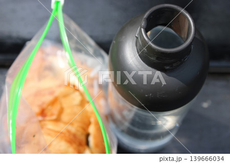 Clear water bottle with a black cap sits beside a ziplock bag filled with sliced fruit, placed on a dark surface illuminated by natural light 139666034
