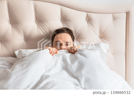 Frightened man lying in bed covering himself with blanket. Concept of fear, anxiety, insomnia and emotional stress. Frightened man lying in bed covering himself with blanket. Concept of fear, anxiety, insomnia and emotional stress. 139666532