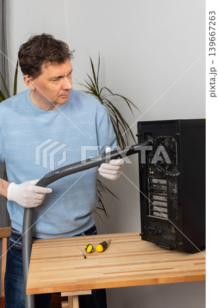 A 53-year-old man cleans dust from a computer system unit with a vacuum cleaner in a tidy room. He carefully removes dirt to keep the device running well A 53-year-old man cleans dust from a computer system unit with a vacuum cleaner in a tidy room. He carefully removes dirt to keep the device running well 139667263