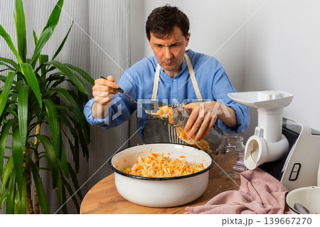 A 53-year-old man works in his kitchen fermenting cabbage and carrots. He carefully adds the mixture from a large bowl into a jar, showing concentration 139667270