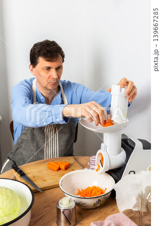 A 53-year-old man is in his kitchen preparing cabbage and carrots for fermentation. He uses a machine to shred the vegetables before placing them in a bowl 139667285