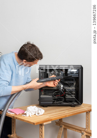 A 53-year-old man uses a vacuum to clean dust from a computer system unit on a wooden table in a home office. The setting is tidy and organized A 53-year-old man uses a vacuum to clean dust from a computer system unit on a wooden table in a home office. The setting is tidy and organized 139667286