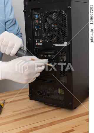 A 53-year-old man removes dust from a computer system unit using a brush while wearing gloves. He is focused on maintaining the computer in a room with plants 139667295