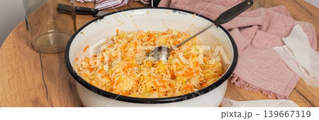 Cabbage and carrots are mixed together for fermentation in a bowl on a wooden table. Jars and kitchen equipment are present in the background, showing a food preparation scene, banner Cabbage and carrots are mixed together for fermentation in a bowl on a wooden table. Jars and kitchen equipment are present in the background, showing a food preparation scene, banner 139667319