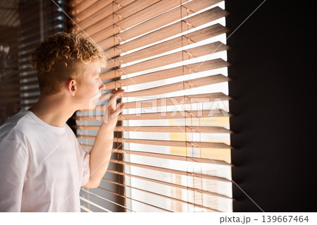 Child observing from window. Boy gazes through blinds quietly Child observing from window. Boy gazes through blinds quietly 139667464
