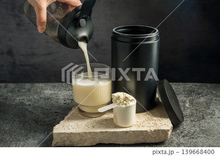 Pouring protein vanilla shake in drinking glass and scoop with whey protein powder on dark background Pouring protein vanilla shake in drinking glass and scoop with whey protein powder on dark background 139668400