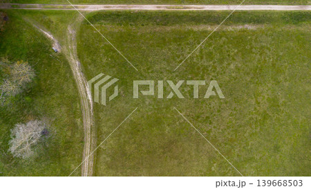 Top down aerial view of a green meadow with a dirt path intersection and a lonely bench. Minimalist rural landscape captured from above, showcasing texture, geometry, and peaceful countryside. 139668503