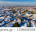 Aerial winter view of a small snowy Dutch town on a sunny day. Red-roofed houses covered in light snow surround a historical church. Clear blue sky and long shadows. 139668508