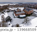 Elevated aerial winter view of the snow-covered Schloss Anholt moat castle in Germany. The historical moated castle is surrounded by ice and snow-laden trees under an overcast sky. 139668509