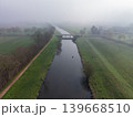 High-angle aerial view of a calm river canal flowing through a misty, foggy agricultural landscape in winter or early spring. A rural road and bridge cross the water, surrounded by bare trees. 139668510