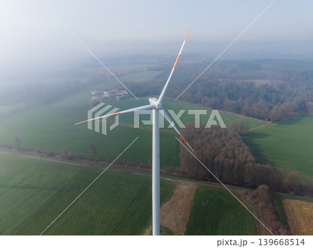 Aerial perspective of multiple wind turbines stretching into a misty, agricultural landscape on a clear, high-altitude morning. Sustainable green energy farm concept. Aerial perspective of multiple wind turbines stretching into a misty, agricultural landscape on a clear, high-altitude morning. Sustainable green energy farm concept. 139668514