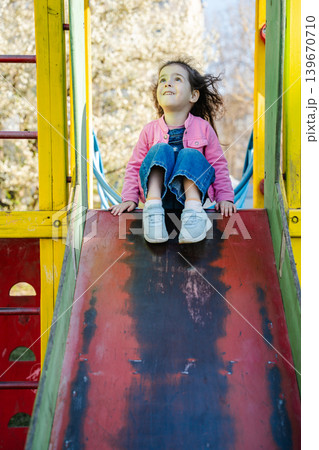 Caucasian girl sitting on playground slide in sunny residential park area. Childhood and outdoor lifestyle 139670710