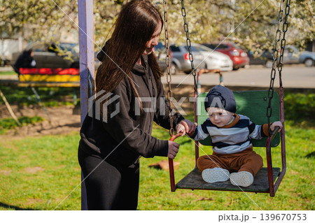 Caucasian mother pushing baby son on playground swing in sunny residential park. Family and parenting lifestyle Caucasian mother pushing baby son on playground swing in sunny residential park. Family and parenting lifestyle 139670753