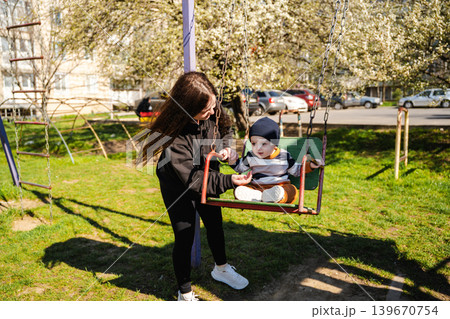 Caucasian mother pushing baby son on playground swing in blooming residential park area. Family and parenting lifestyle Caucasian mother pushing baby son on playground swing in blooming residential park area. Family and parenting lifestyle 139670754