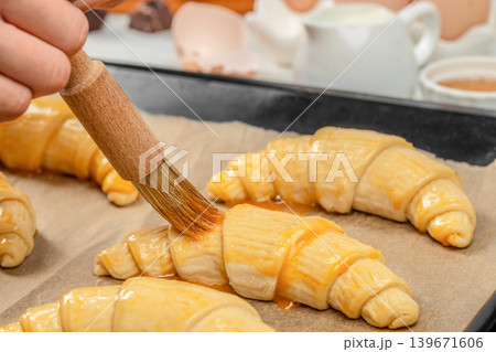 Applying egg wash on raw croissants before baking, close up pastry preparation process Applying egg wash on raw croissants before baking, close up pastry preparation process 139671606