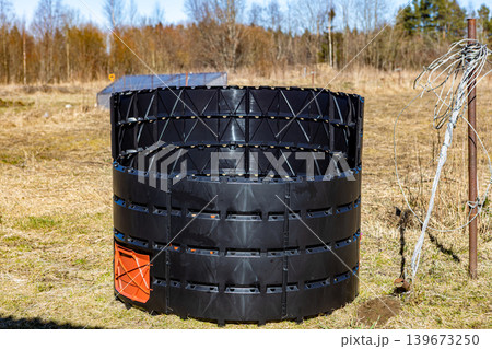 Black circular compost bin with orange door, made of plastic panels, standing outdoors on grass, ready for organic waste processing. Black circular compost bin with orange door, made of plastic panels, standing outdoors on grass, ready for organic waste processing. 139673250