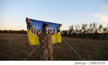 Young male military in uniform standing with raised flag Ukraine above his head at field. Soldier of ukrainian army lifted blue-yellow banner as symbol of victory against russian aggression on field Young male military in uniform standing with raised flag Ukraine above his head at field. Soldier of ukrainian army lifted blue-yellow banner as symbol of victory against russian aggression on field 139676297