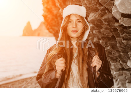 Woman beach sunset Young woman wearing a leather jacket and winter hat poses against a rocky cliff by the sea at sunset 139676963