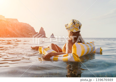 Woman, Beach, Relaxation: A woman in a hat floats on an inflatable raft in the ocean, enjoying the view of a rocky coastline. Woman, Beach, Relaxation: A woman in a hat floats on an inflatable raft in the ocean, enjoying the view of a rocky coastline. 139676989