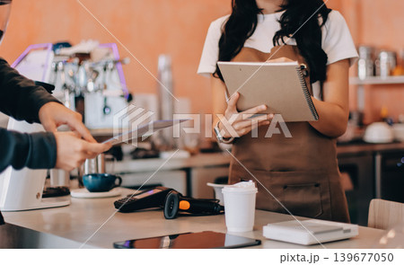 Friendly Asian female barista receives a credit card from a customer for payment. Both women are smiling in a warmly lit coffee shop with a rustic interior. Cashless payments. Small family business Friendly Asian female barista receives a credit card from a customer for payment. Both women are smiling in a warmly lit coffee shop with a rustic interior. Cashless payments. Small family business 139677050